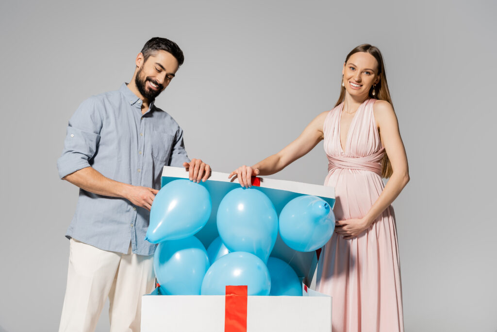 smiling and elegant pregnant woman opening big gift box with blue balloons near husband during baby shower isolated on grey, expecting parents concept, gender party, it`s a boy smiling and elegant pregnant woman opening big gift box with blue balloons near husband during baby shower isolated on grey, expecting parents concept, gender party, it`s a boy